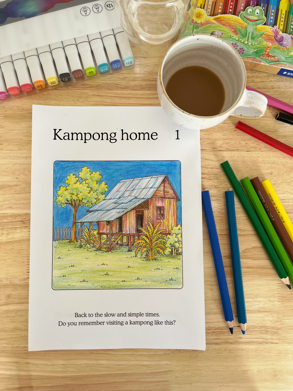 Coloring page with a kampong illustration on a table with coloring supplies